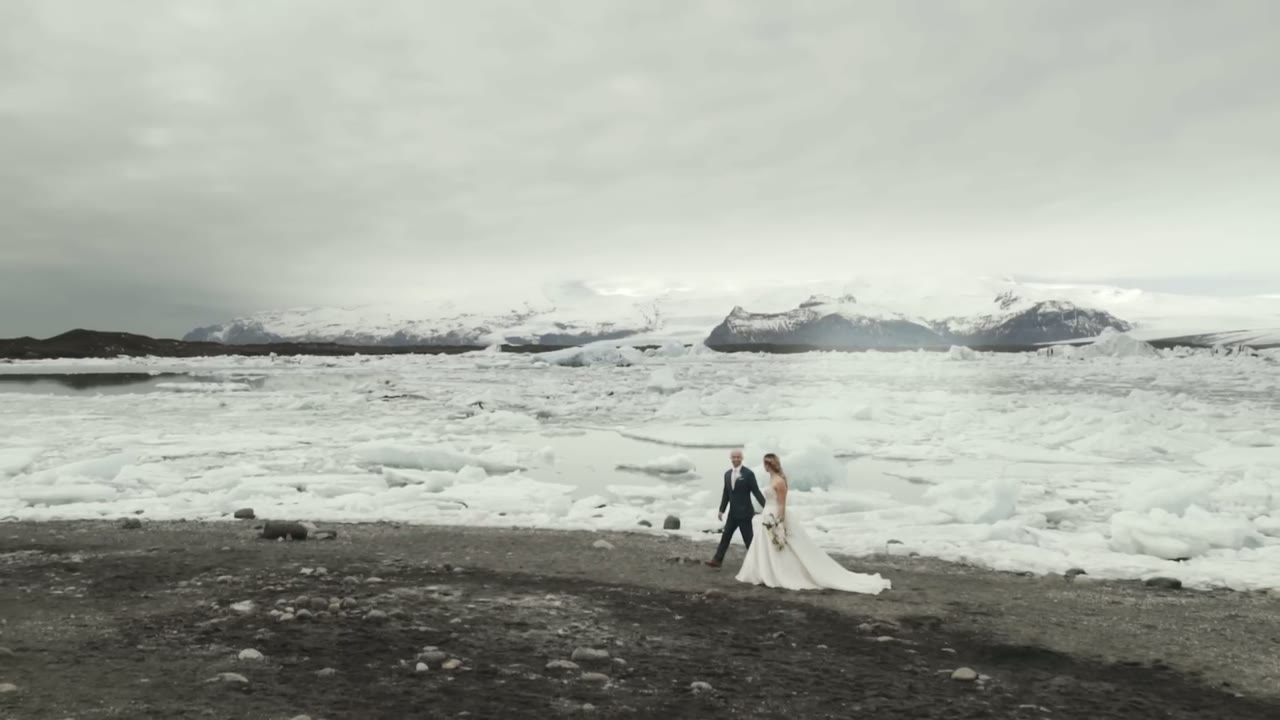 Aerial view of the bride and groom walking tiny across a black volcanic beach beside floating icebergs