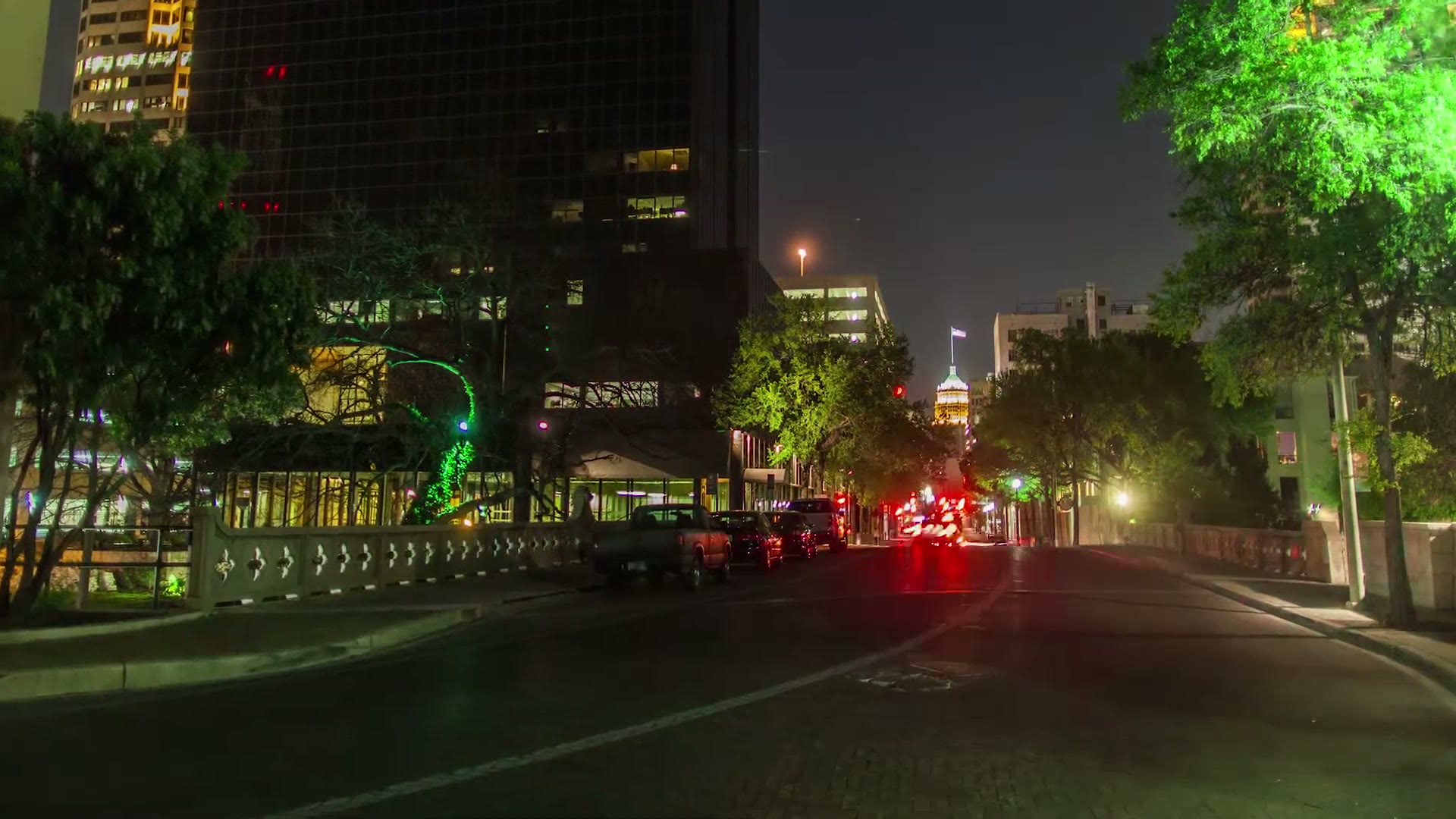 A downtown San Antonio street at night with lit trees, traffic lights and a tower in the distance