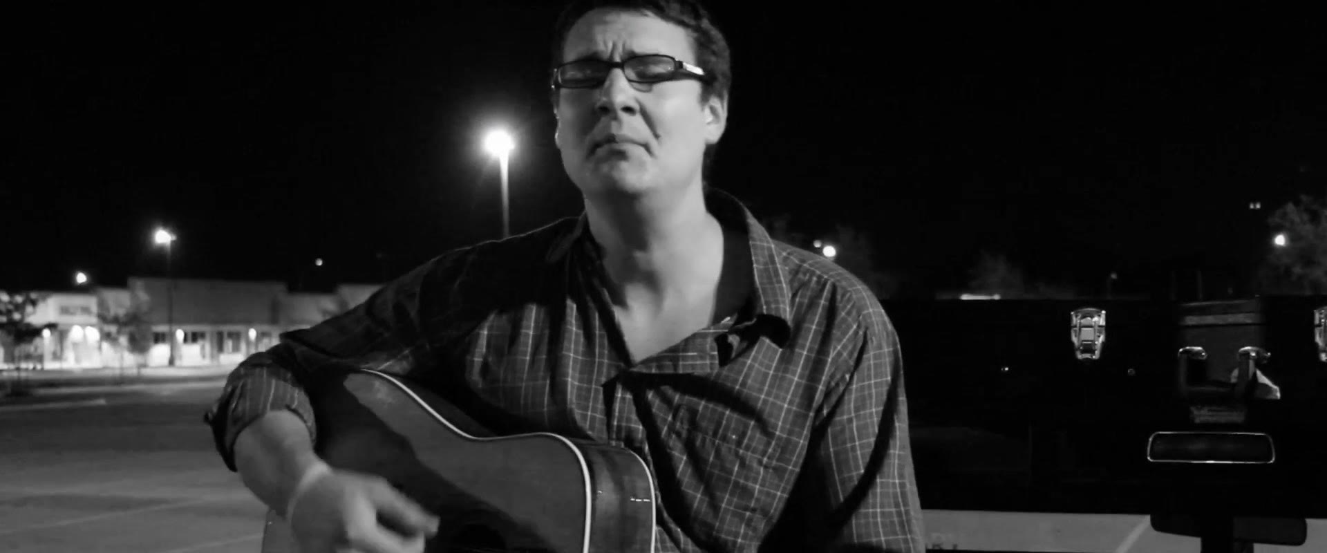 Black and white shot of the musician singing with his head tilted back and eyes closed, guitar in hand in an empty parking lot at night