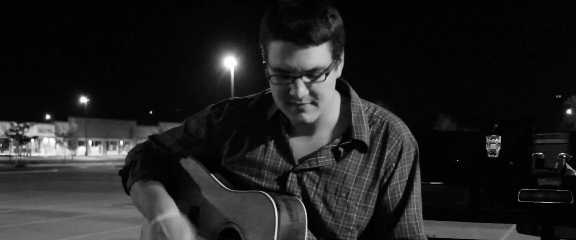 Black and white shot of the same musician looking down at his guitar with a faint smile while playing