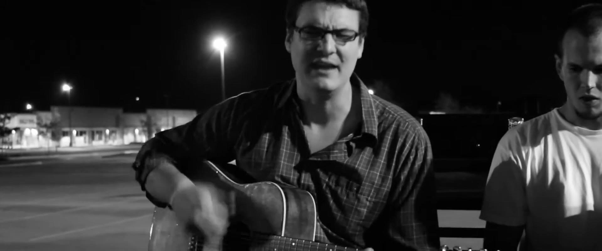 Black and white shot of Curt Vernon singing and playing acoustic guitar in an empty parking lot, a friend listening at right
