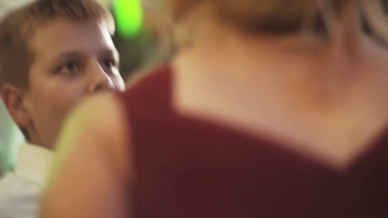 A young boy watching a woman in a maroon dress dancing at the reception, close and blurred