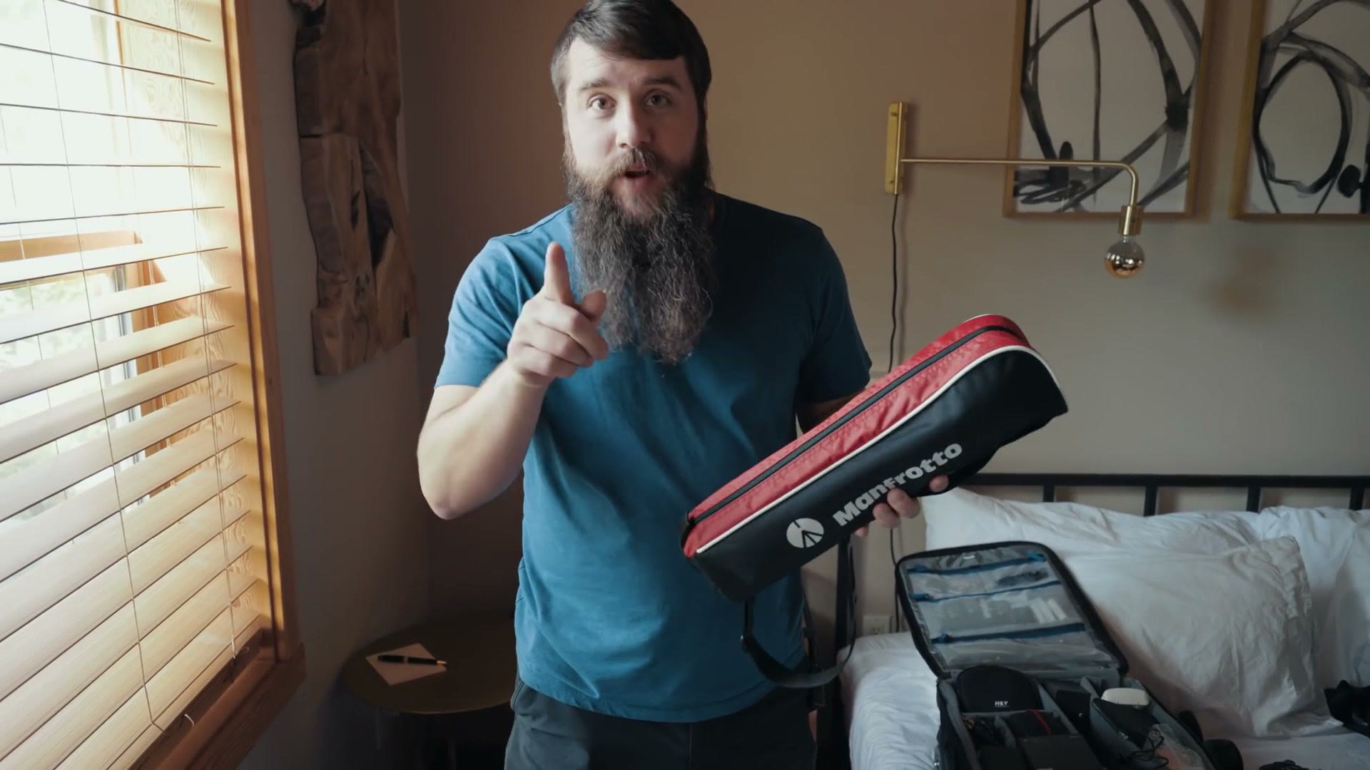 Matt holding a compact Manfrotto tripod case in a hotel room, with an open camera bag on the bed behind him.