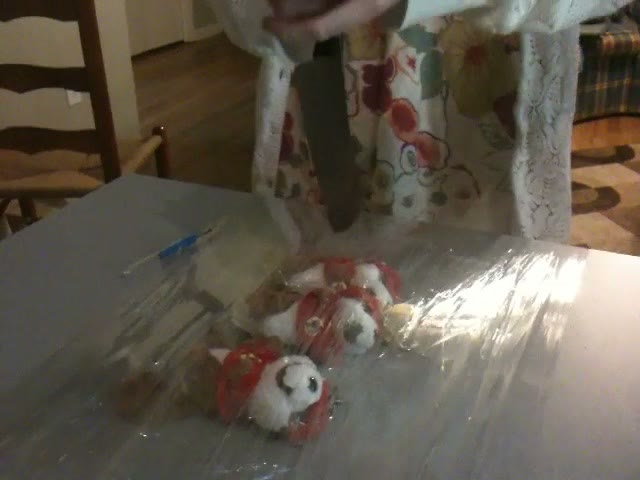 A dining table covered in plastic sheeting with several plush beaver toys wrapped in cling film and a knife lying beside them