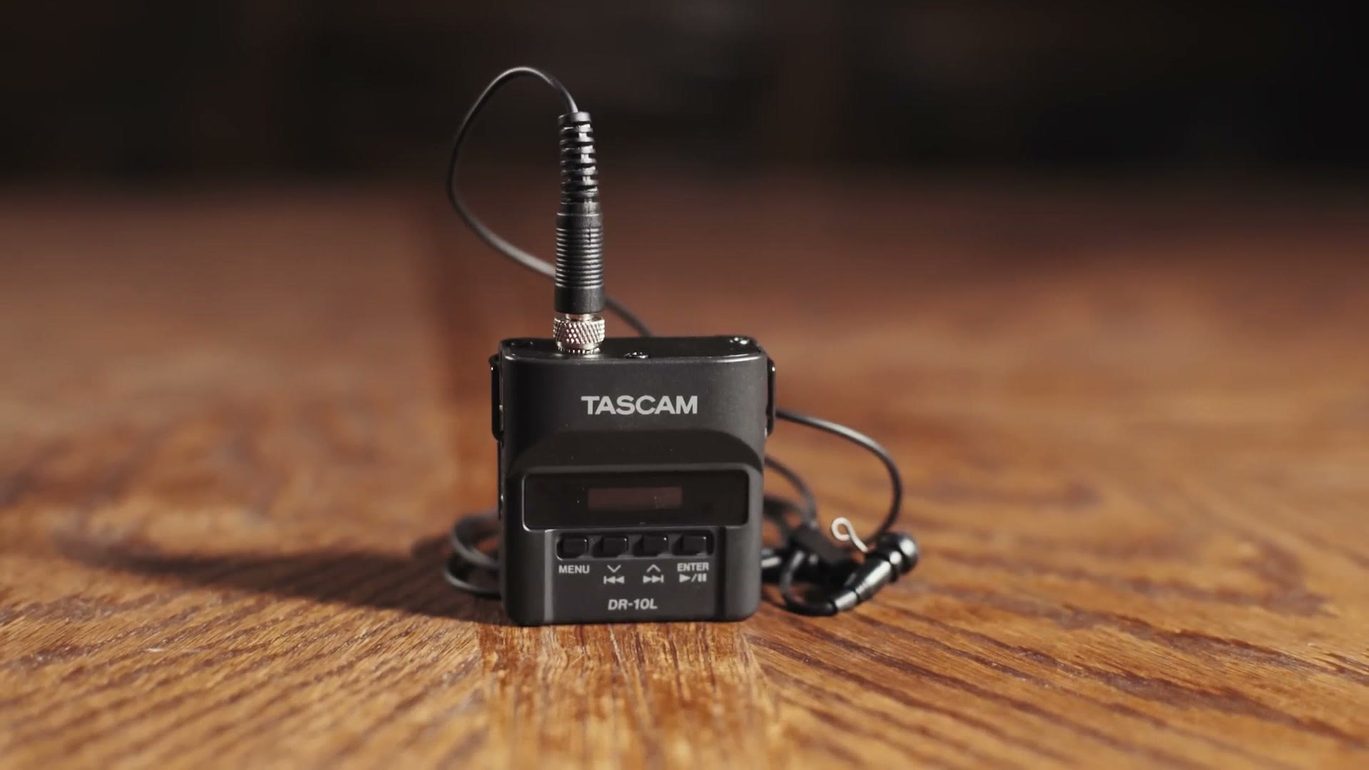 A black Tascam DR-10L audio recorder standing on a wooden table with its lavalier mic and cable coiled beside it.