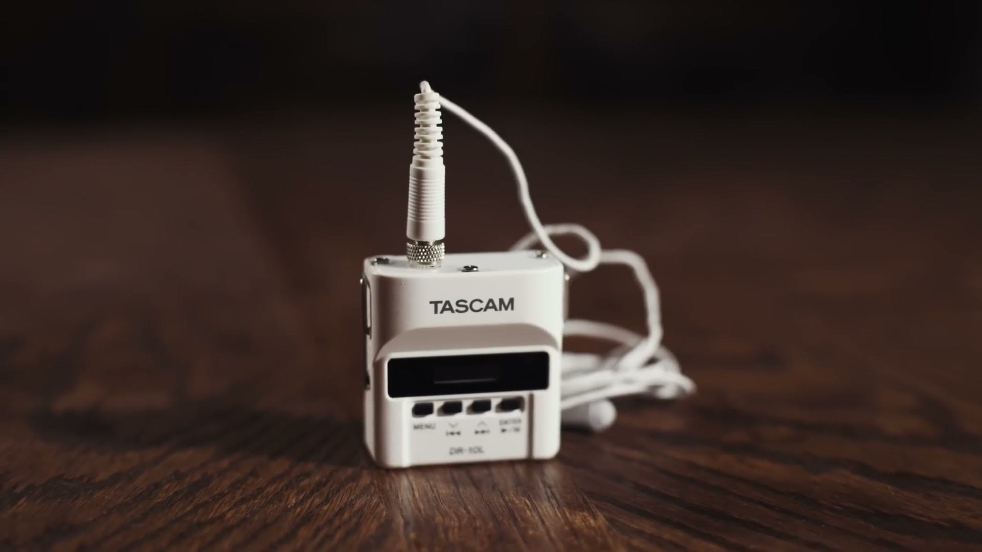 A white Tascam DR-10L recorder with a white lavalier mic and white cable, standing on a wooden table.