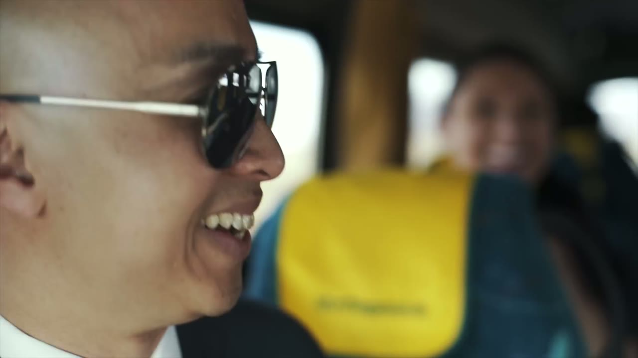 Bald man wearing sunglasses laughing inside a bus with yellow and teal seats, a woman smiling blurred behind him