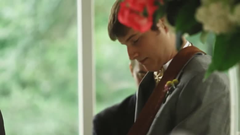 A guitarist in a grey suit playing during the wedding, seen through out-of-focus red and white flowers in the foreground