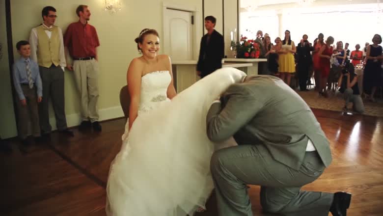 The groom kneeling in front of the seated bride during the garter removal while the guests watch from around the dance floor
