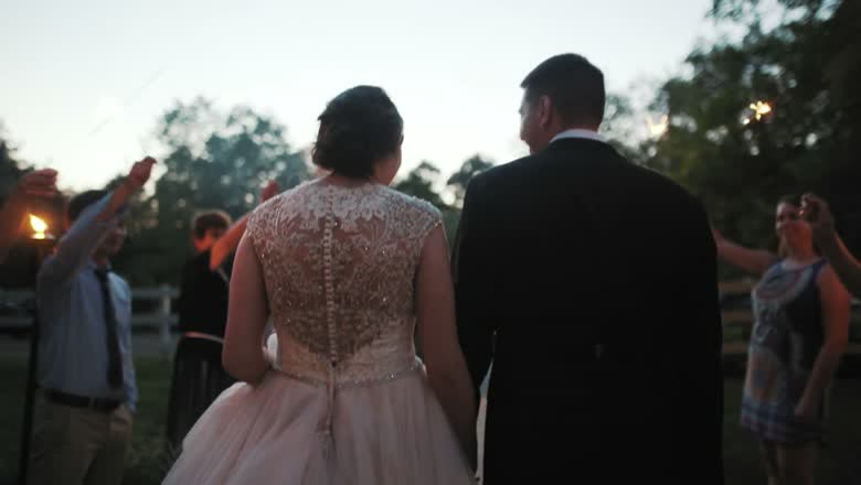 The couple walking out hand in hand at dusk through a short line of guests holding sparklers, with trees behind