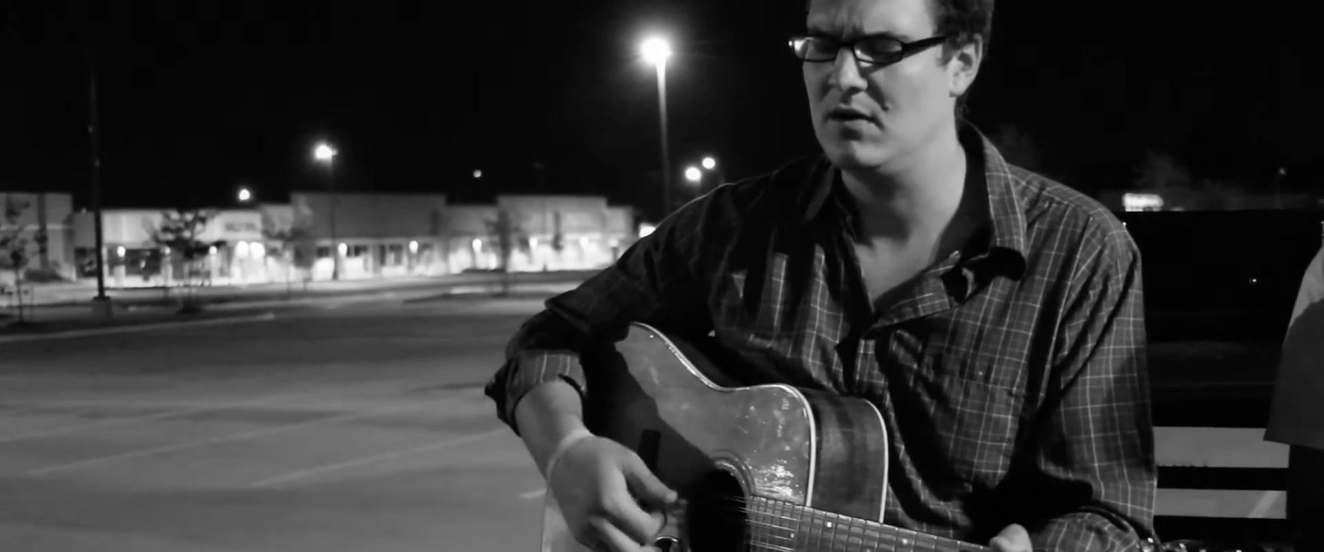Wider black and white shot showing the empty lot and a tall single streetlight beside lit storefronts as Curt Vernon plays