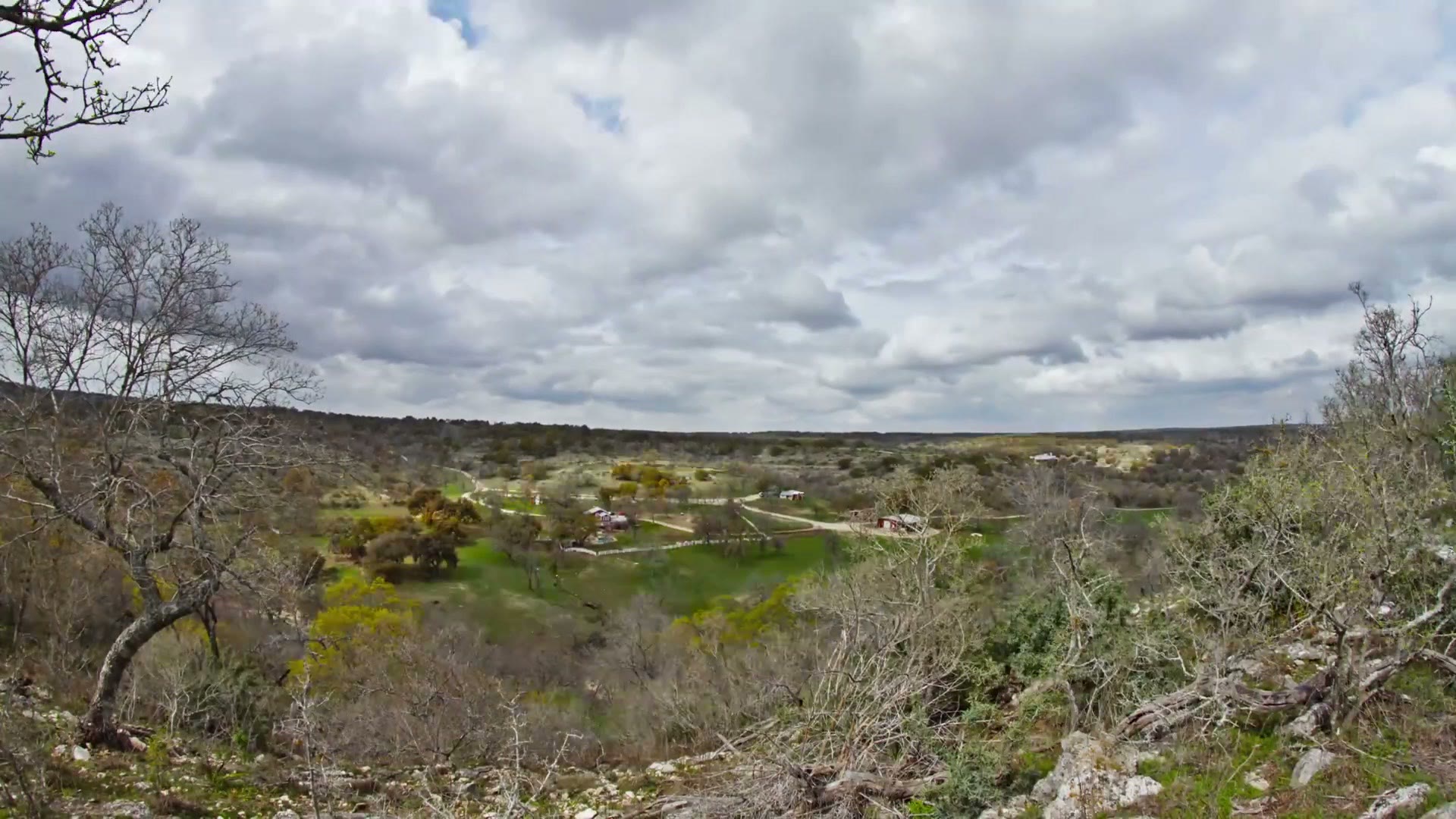 Wide timelapse frame looking down over a Texas ranch valley, with bare winter trees in the foreground and a thick overcast sky above