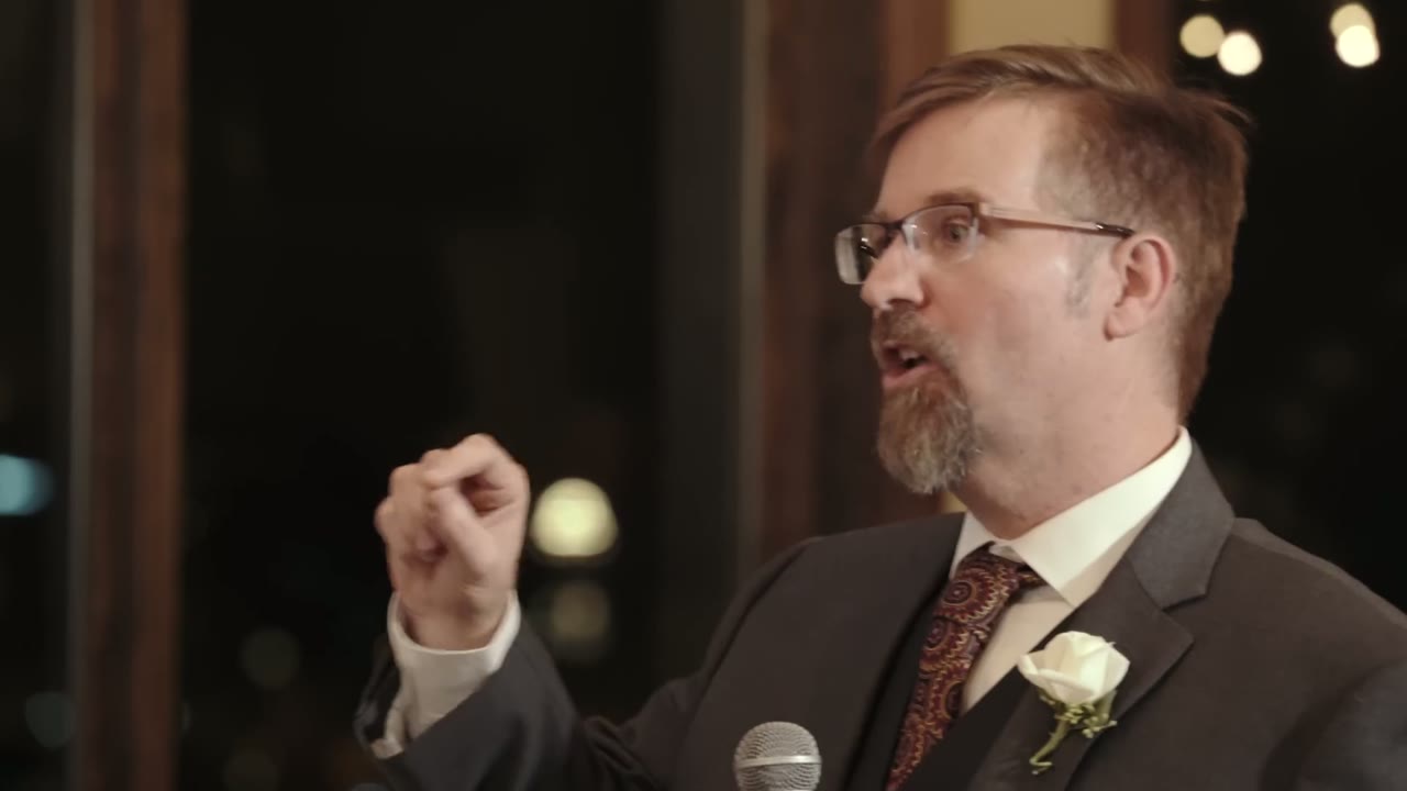A man in glasses giving a toast at the reception, gesturing with a raised fist