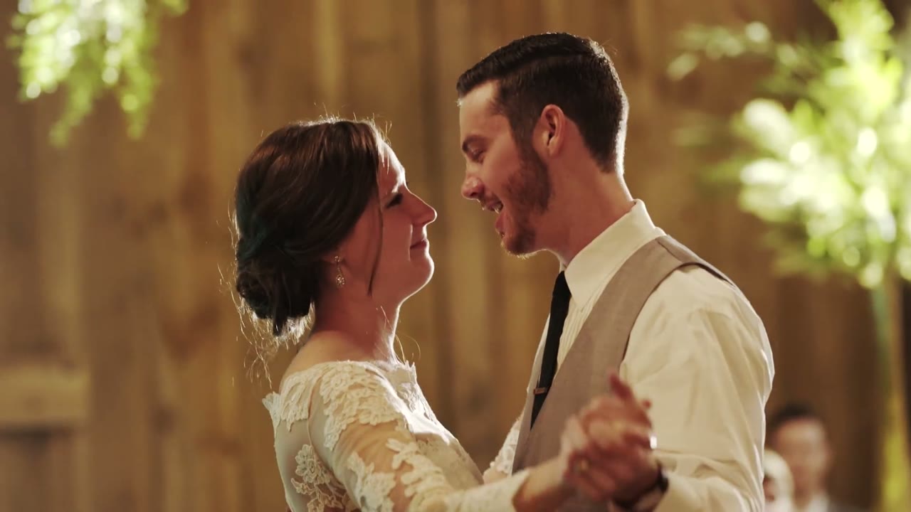 Bride and groom slow dancing in a wood-paneled barn, foreheads close, warm greenery lit behind them