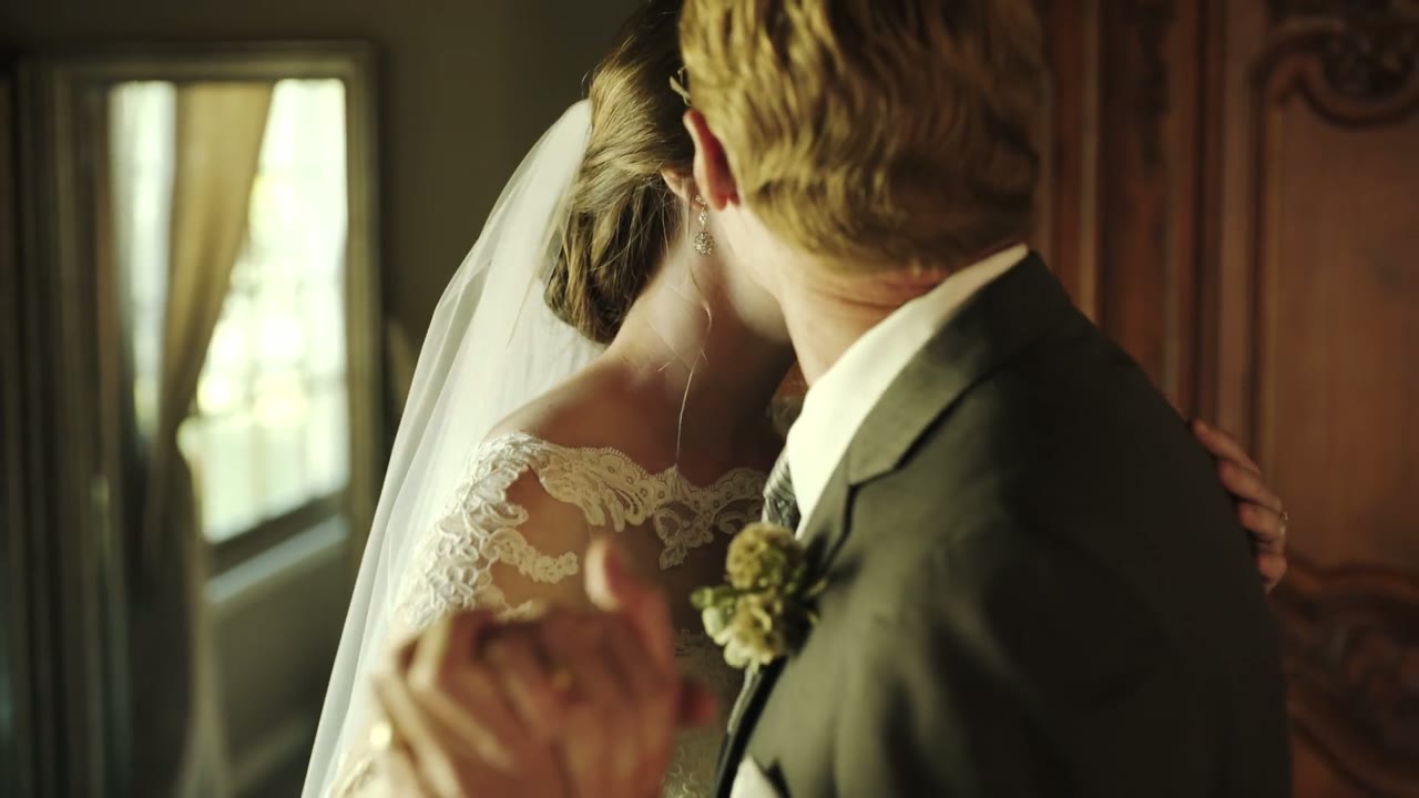 Bride and groom embracing from behind, veil and lace sleeve visible, in a wood-paneled room with a mirror
