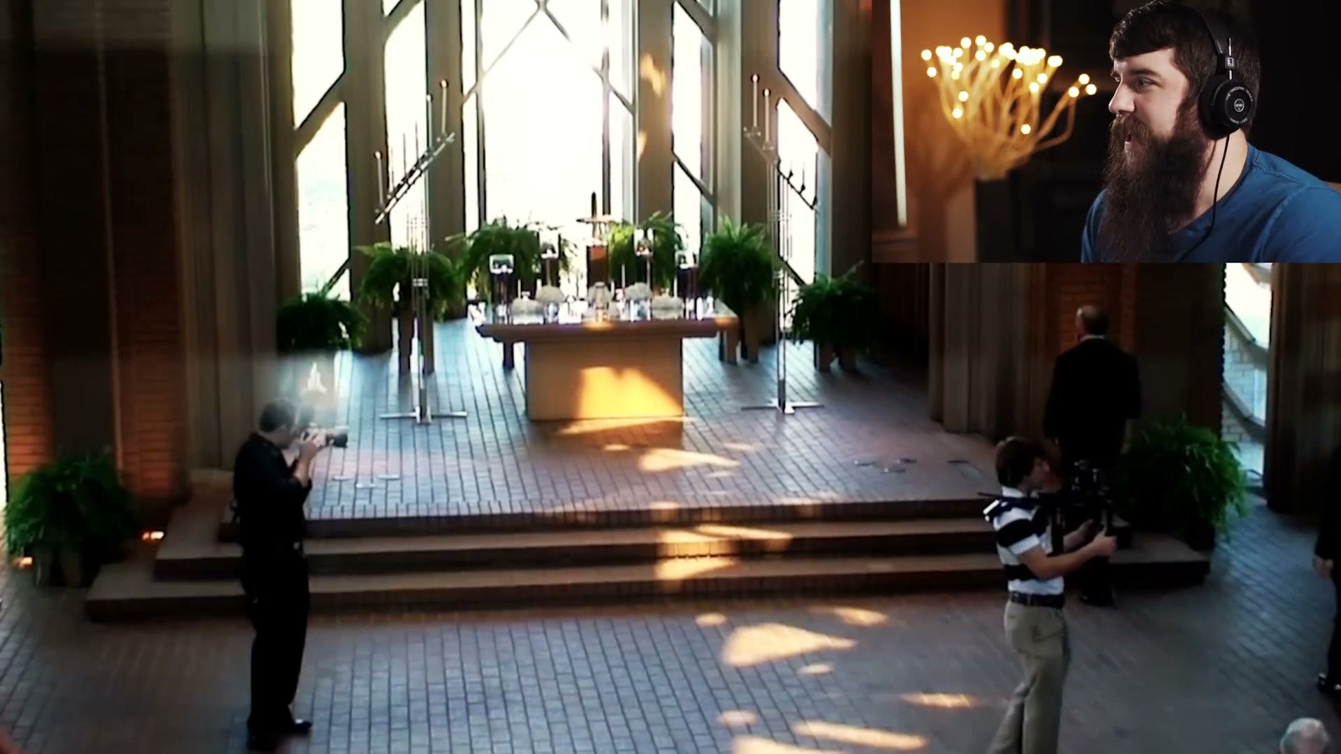 A balcony view of a chapel showing a young Matt in a striped shirt and khakis operating a camera on a heavy shoulder rig while the photographer works nearby.