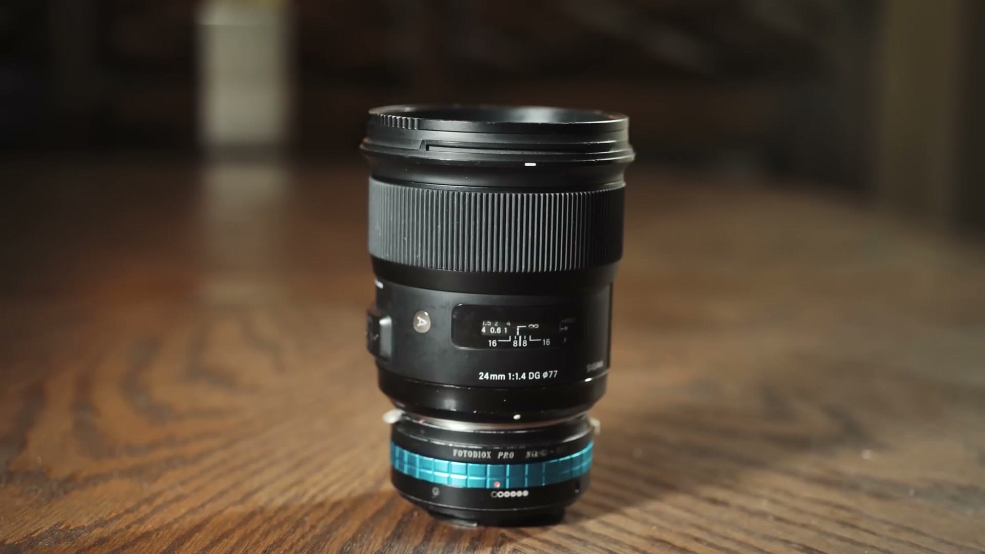 A Sigma 24mm f1.4 DG lens standing on a wood table with a blue ringed Fotodiox Pro lens adapter attached to the mount
