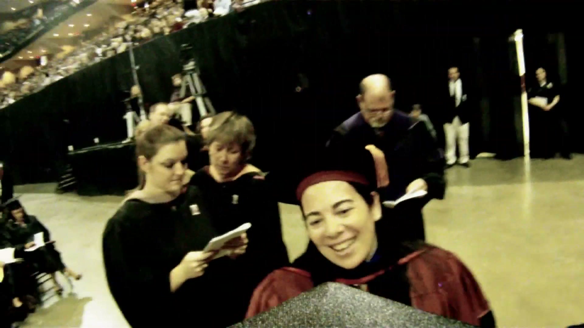 Faculty members in academic regalia at the side of the stage looking directly at the camera on the cap and smiling