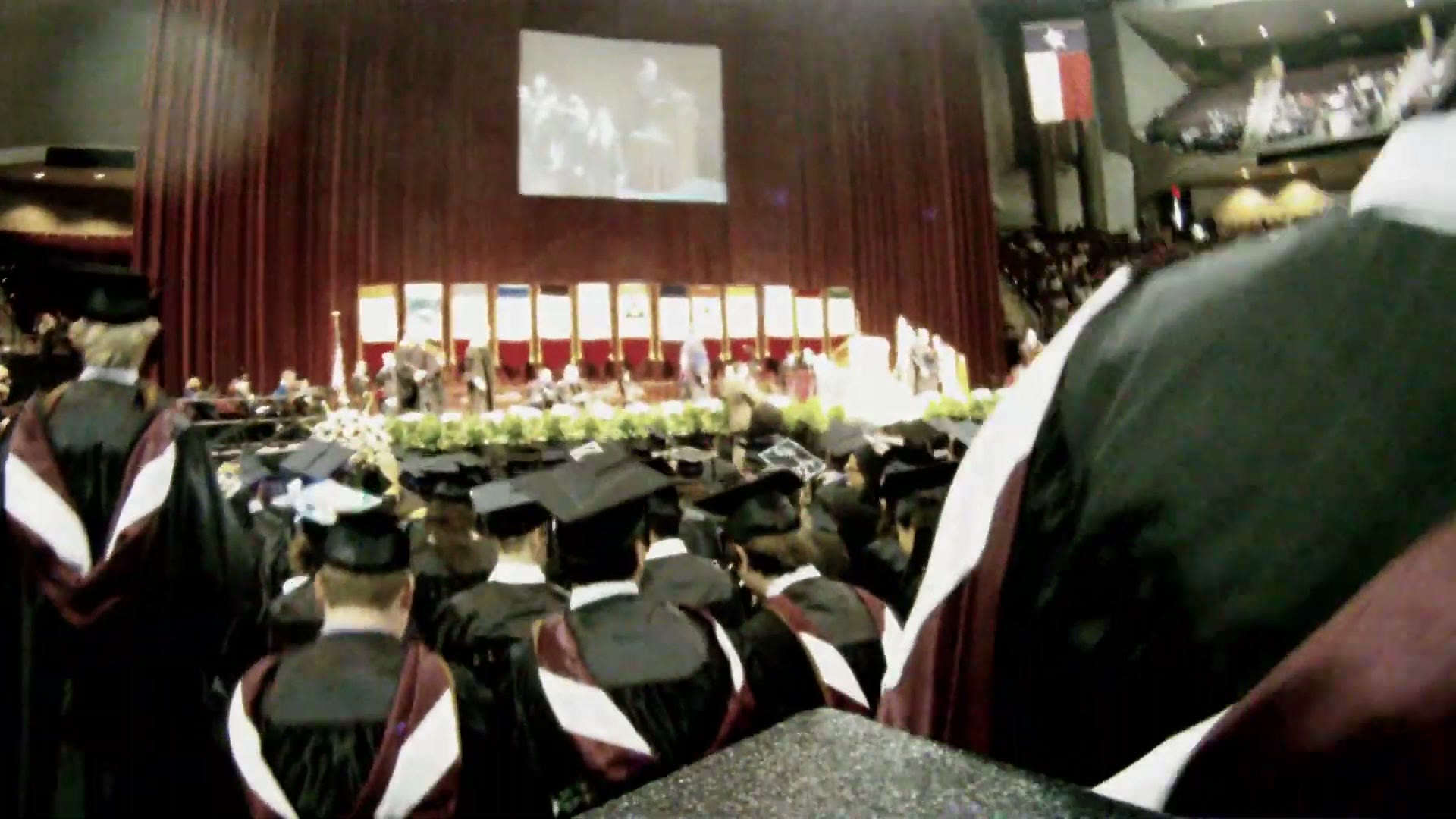 A first person view from a graduation cap looking across rows of seated graduates in black gowns toward a lit stage with a large projection screen