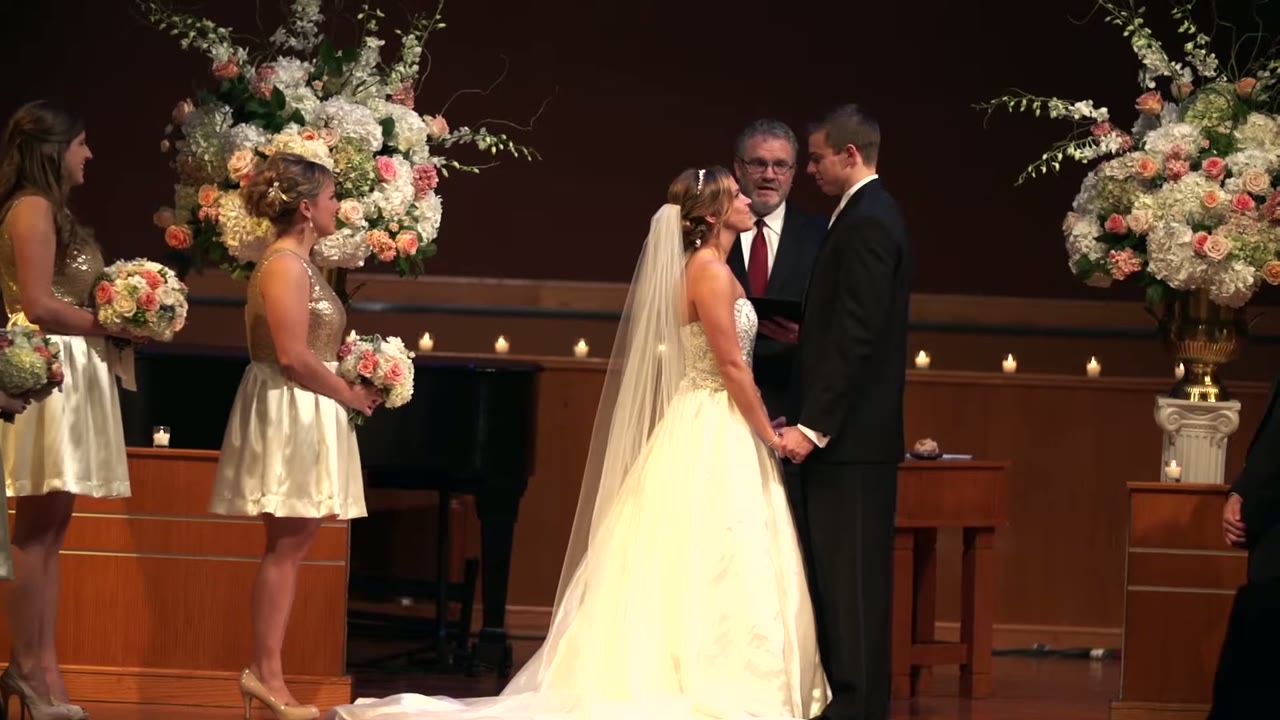 Bride and groom holding hands at the altar with the officiant, tall white and pink floral arrangements on either side