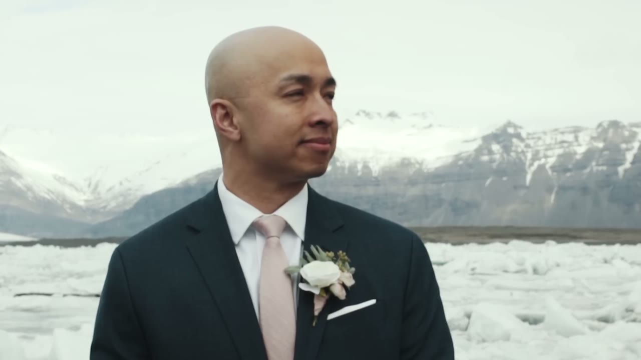 Groom in a navy suit and pink tie standing before the glacier lagoon, snow-covered mountains behind him