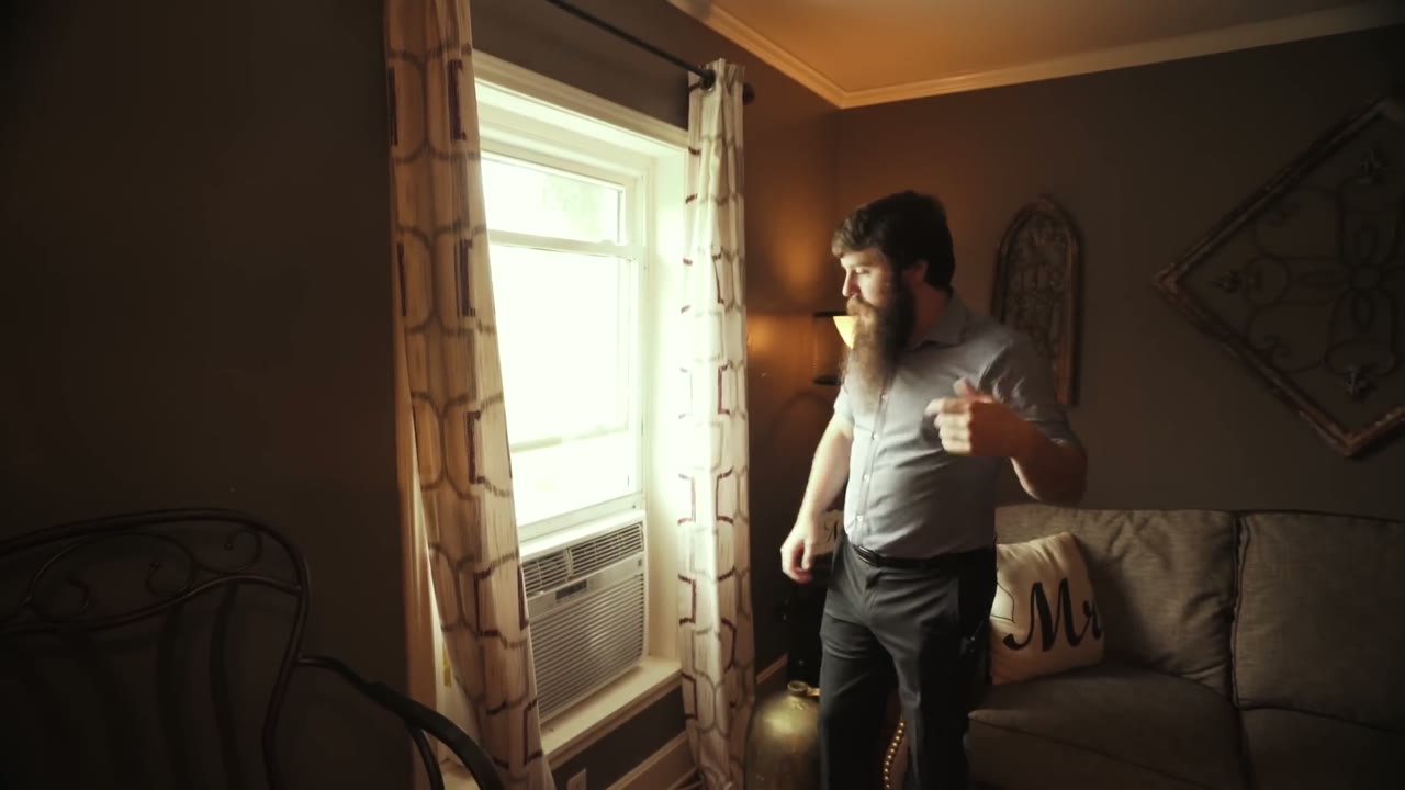 Matt standing near a dim curtained window with a wall AC unit, a gray couch with an Mr pillow behind him