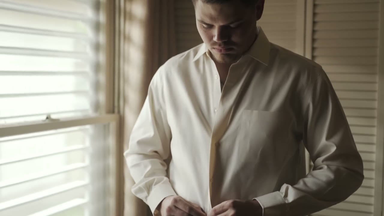 Groom buttoning his white dress shirt cuffs, standing beside a sunlit window with blinds