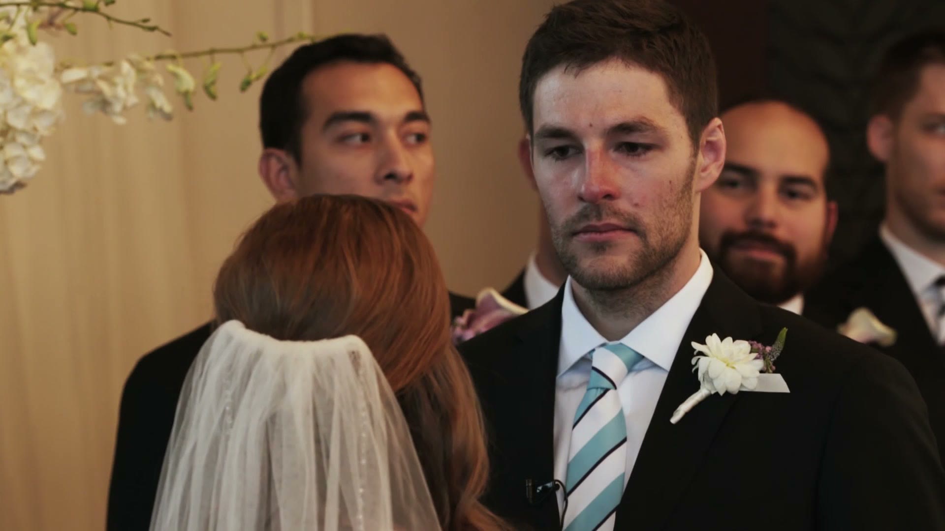 Groom looking emotional as the bride faces him at the altar, groomsmen and a white floral arch behind