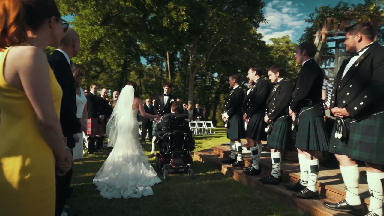Bride being walked down the outdoor aisle past groomsmen in kilts, one guest in an electric wheelchair beside her