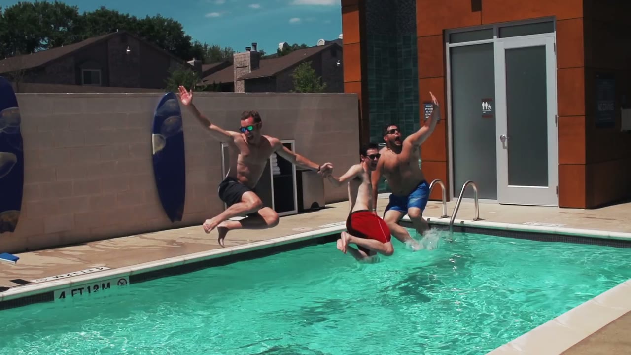 Groomsmen jumping into a pool mid-air in their swim trunks, sunglasses and drinks in hand