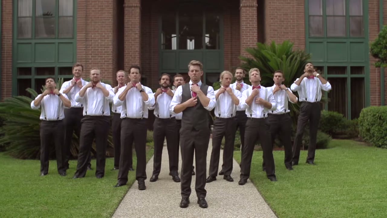 Groom and eleven groomsmen adjusting their pink bow ties in unison on a brick building's front walk