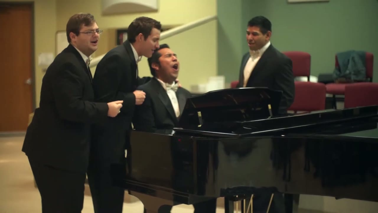 Four groomsmen in tuxedos singing together around a grand piano in a hallway