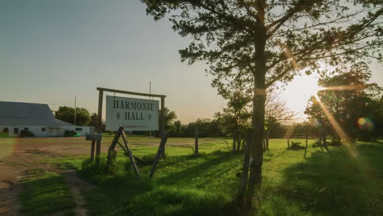 The Harmonie Hall sign at the end of a gravel drive in open Texas countryside, with low sun through the trees behind it