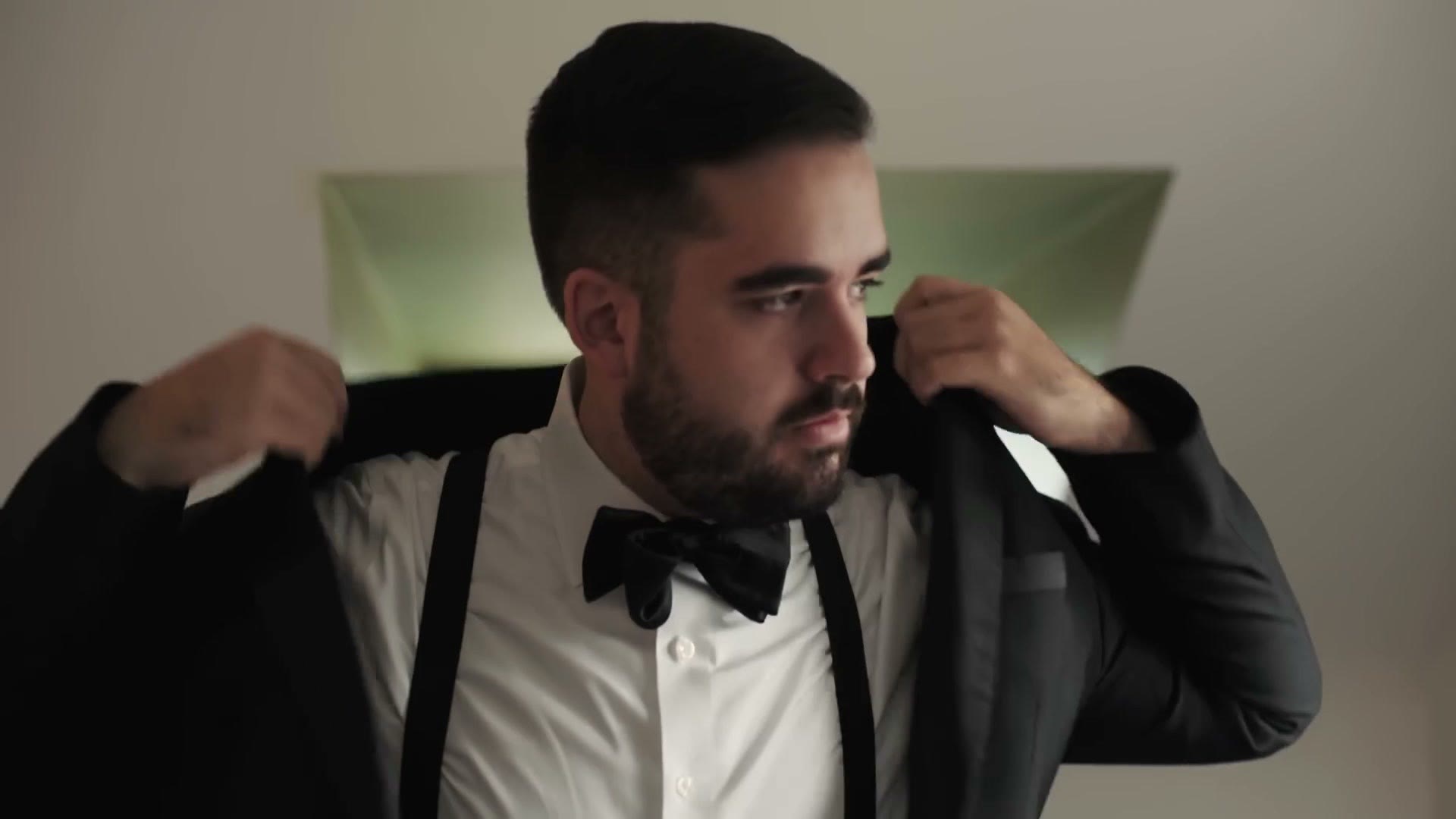 A groom in a tuxedo and bow tie pulling on his jacket in a bright room