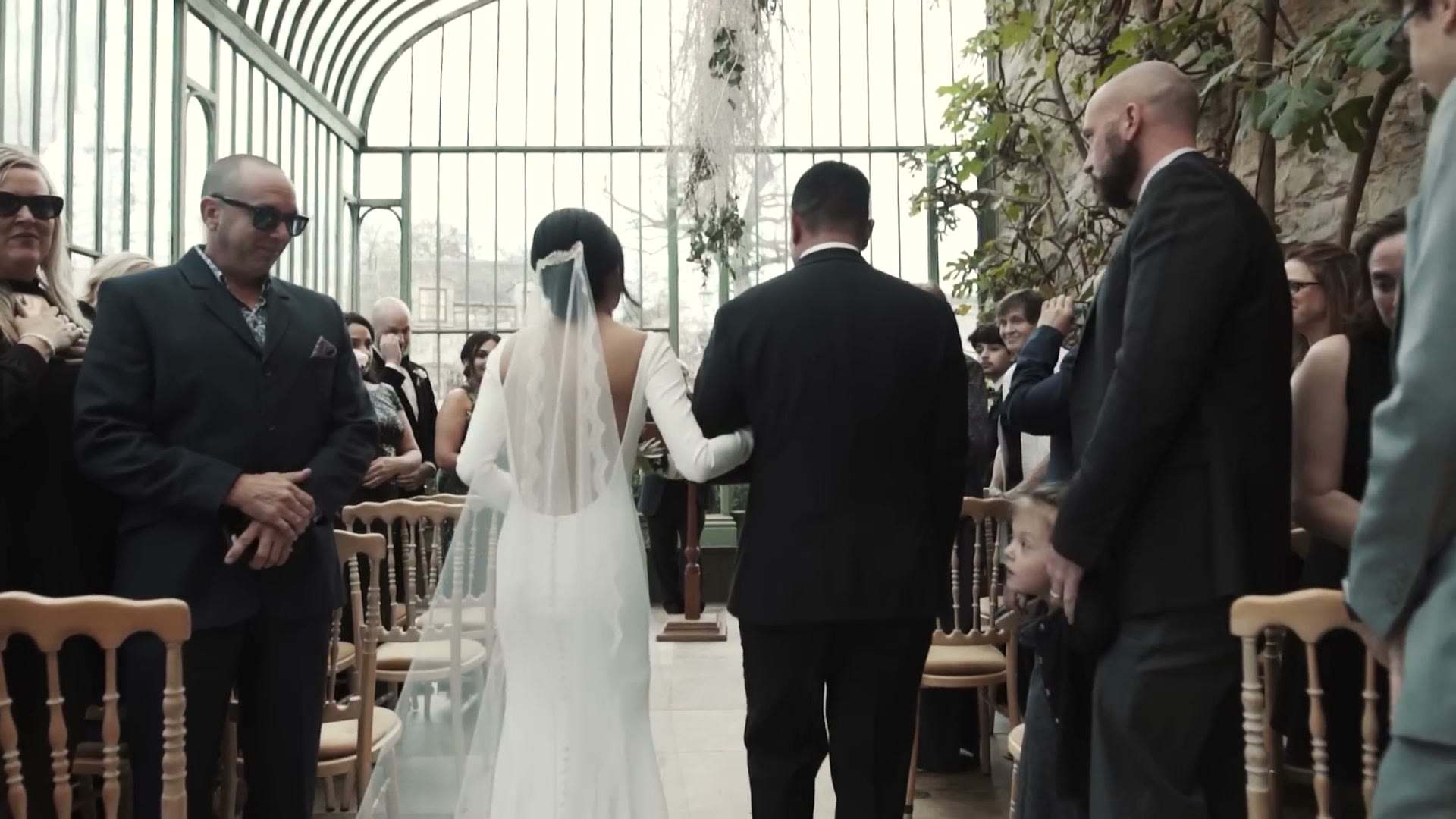 A bride walking down the aisle on her father's arm in a glasshouse ceremony space filled with plants and guests
