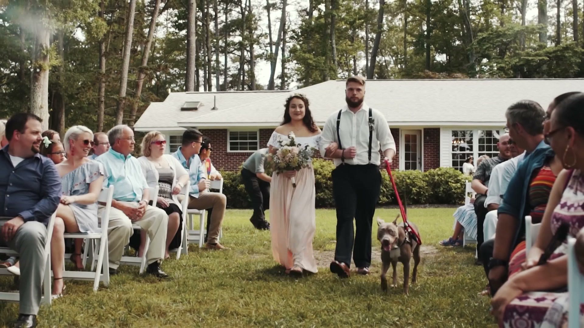 A bride and groom walking back down the aisle at an outdoor backyard wedding, leading a dog on a leash past seated guests