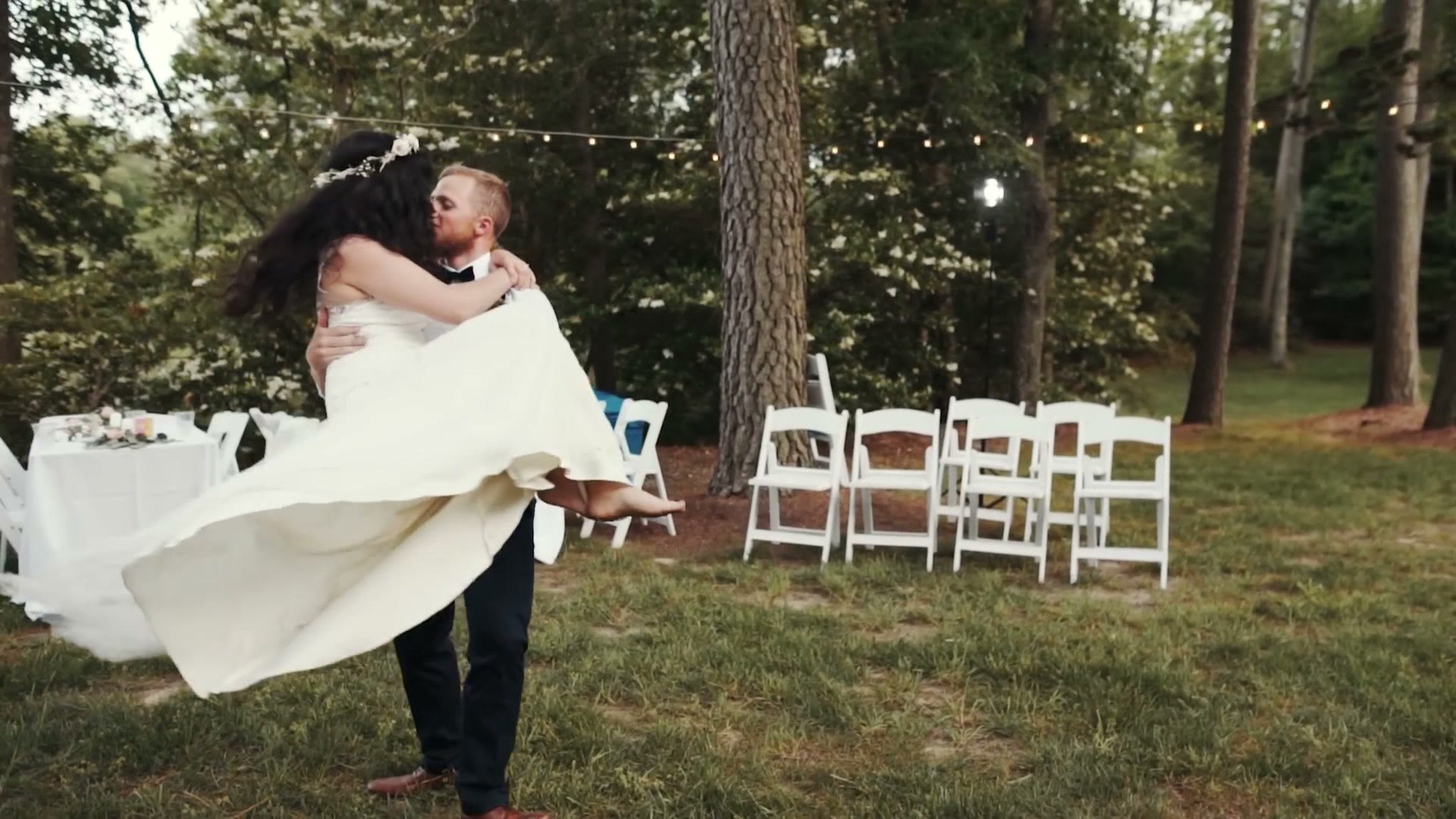 A groom lifting his bride and spinning her under trees hung with string lights at an outdoor wedding