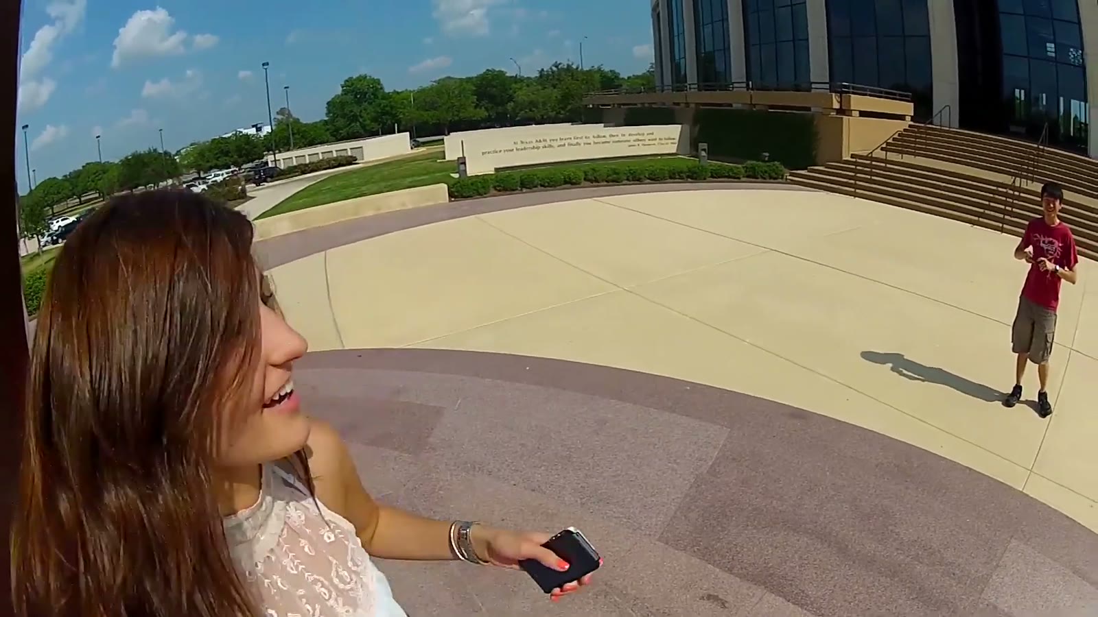 Fisheye selfie of a woman laughing while holding a phone, a man visible in the plaza behind her near a building with steps