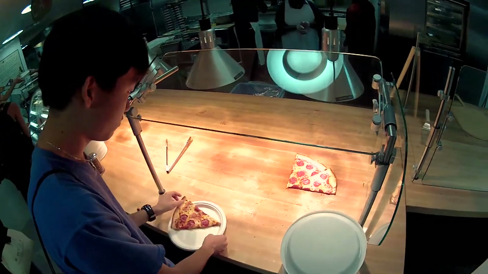 Young man eating a slice of pizza at a wooden table under heat lamps in the dining hall kitchen