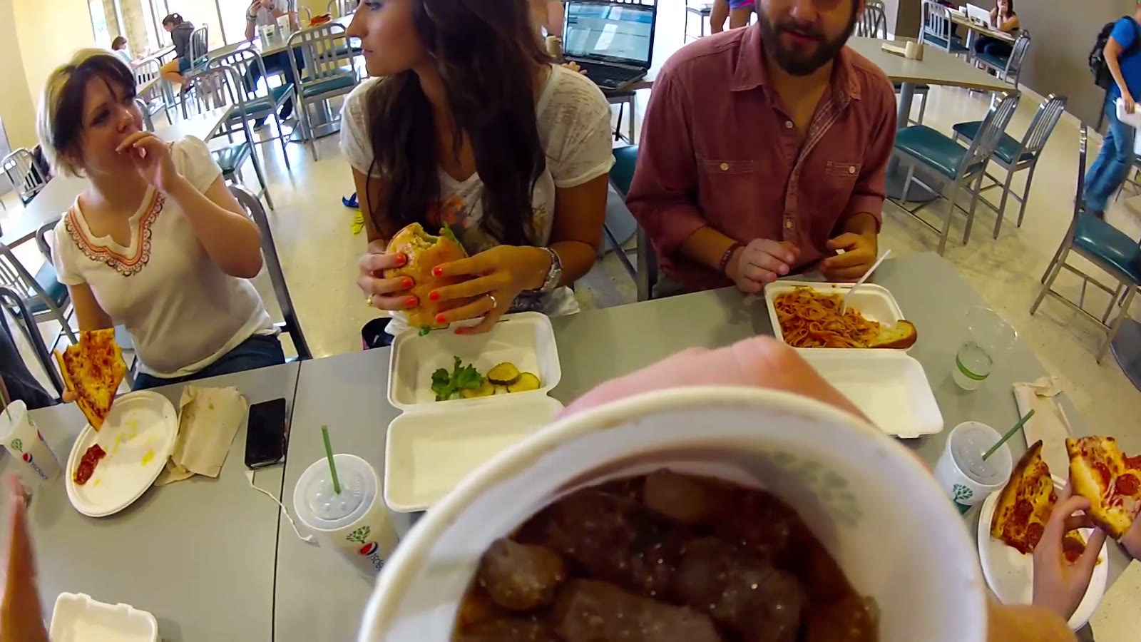 Fisheye view of a dining hall table with a hand holding a bowl of meatballs in the foreground and two women and a man eating behind it