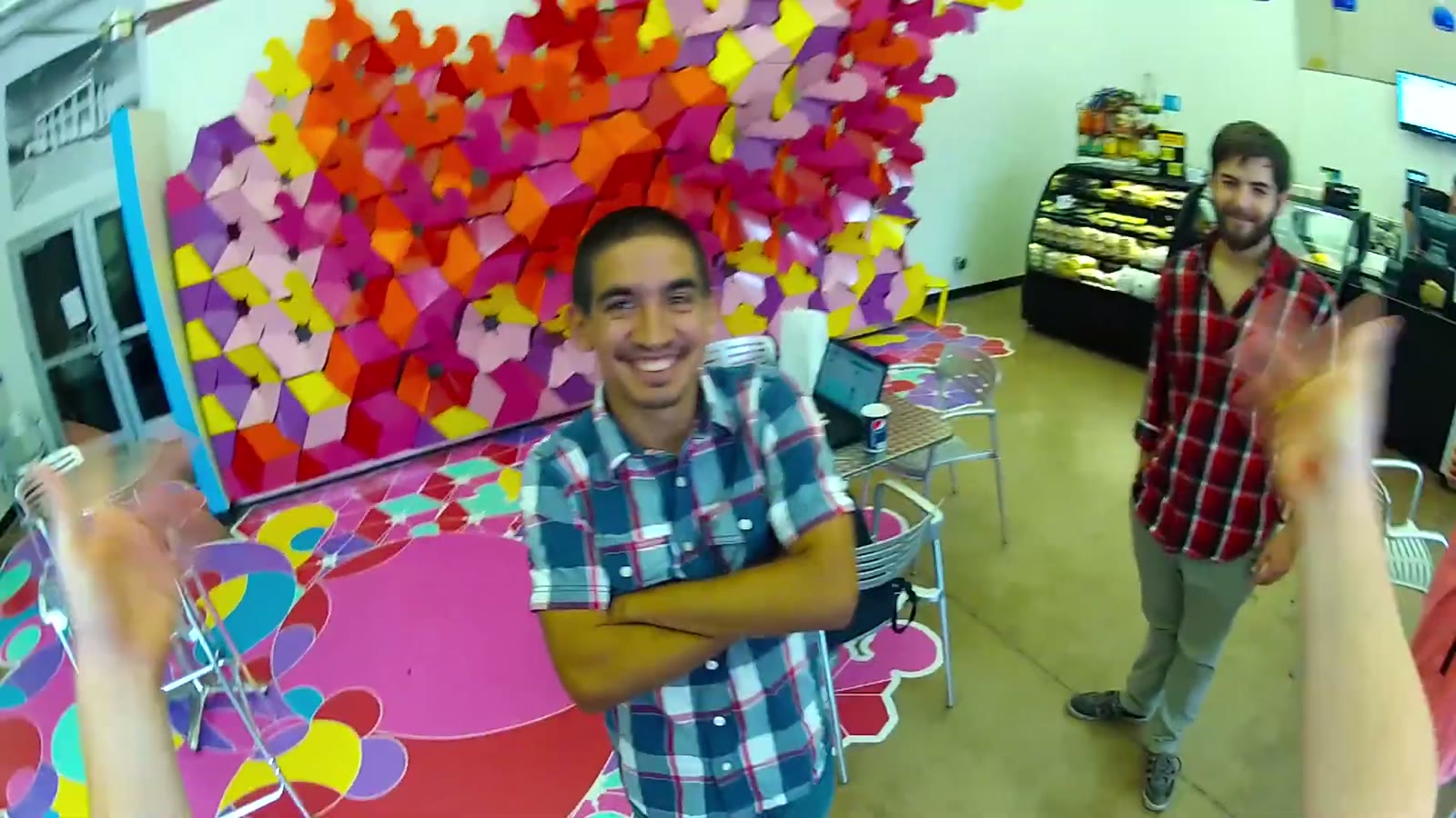 Fisheye selfie of a man laughing with arms crossed in a colorful lounge with geometric wall art, another man waving behind him