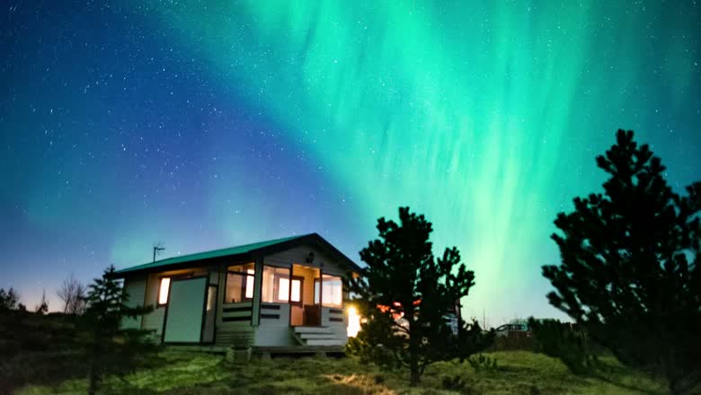 A small lit cabin at night under a full green aurora filling the sky above the trees