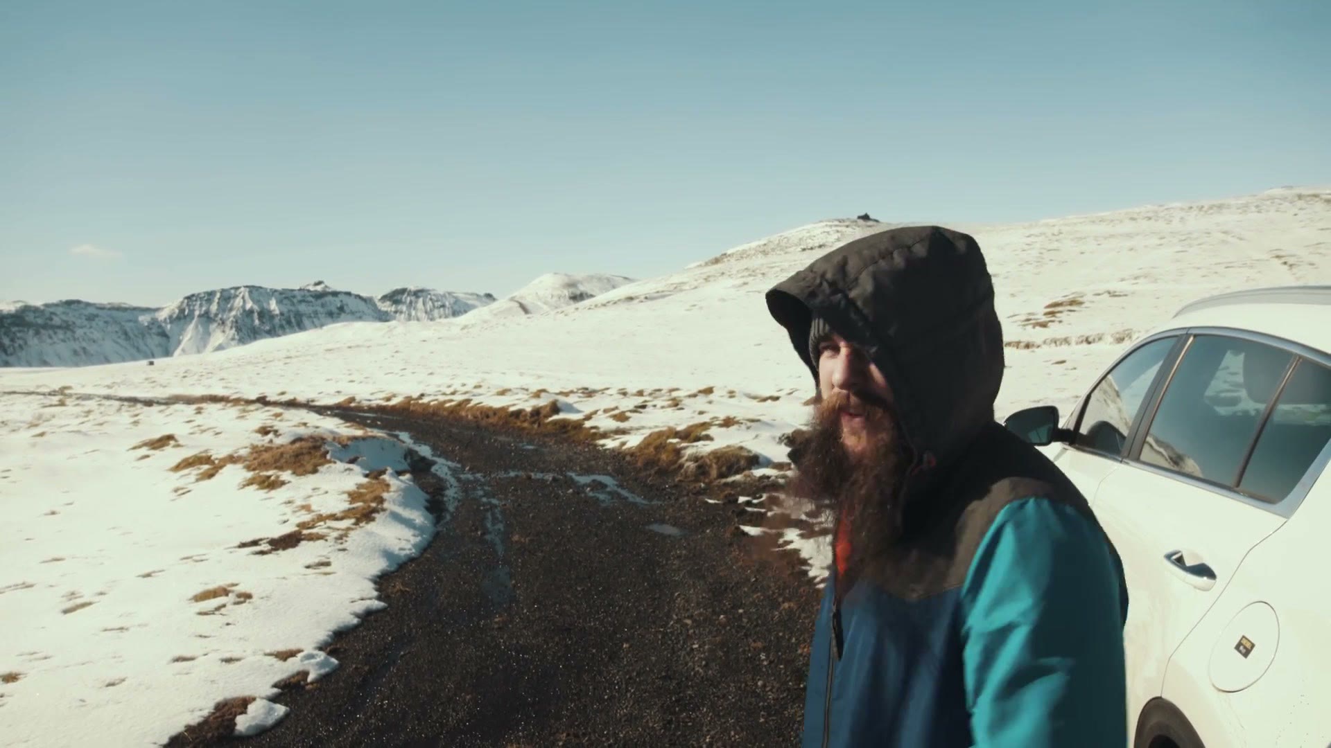 Matt in a hooded winter coat standing beside a white rental car on a snow covered Icelandic road with mountains behind