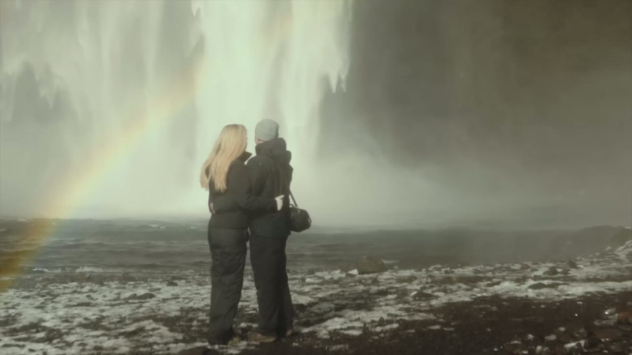 Couple hugging with their backs to camera facing a massive waterfall, a rainbow arcing beside them
