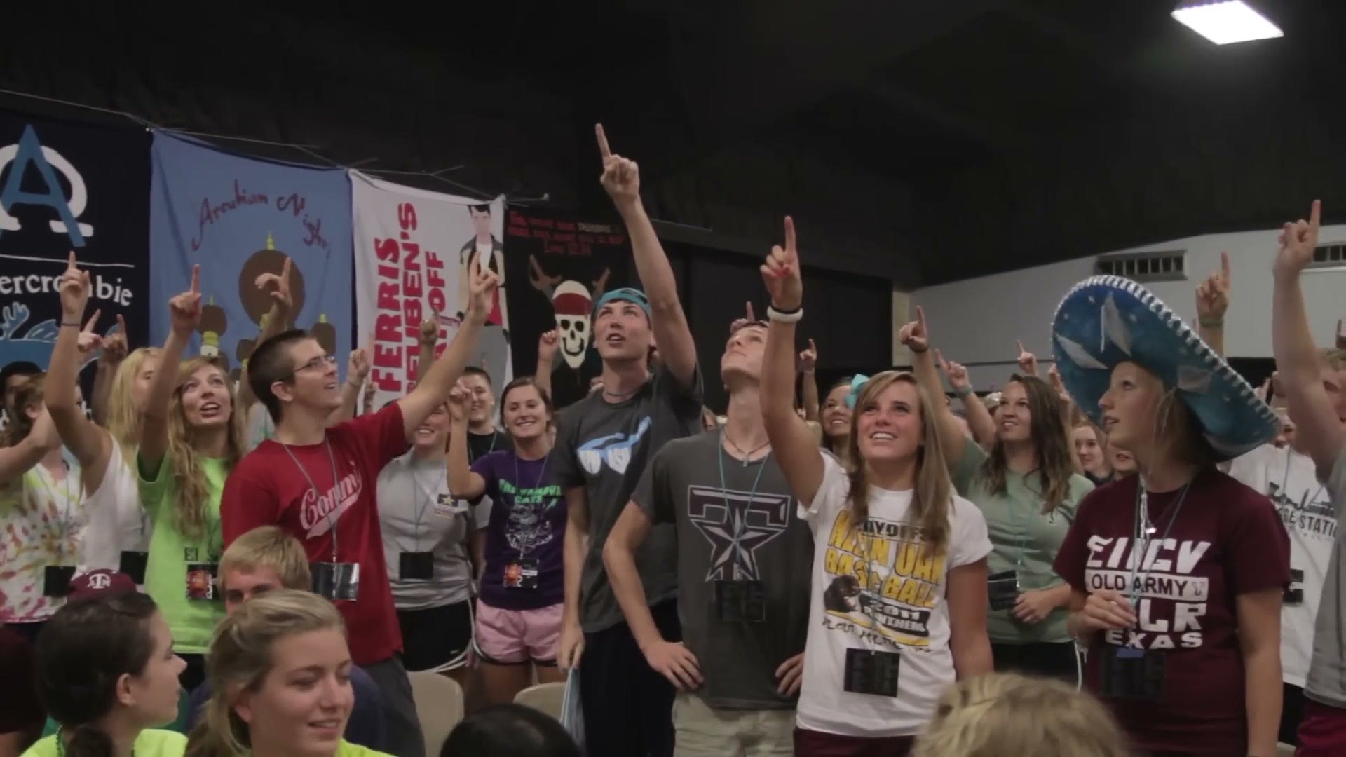 A packed camp meeting hall full of students with hand painted team banners hanging on the wall behind them