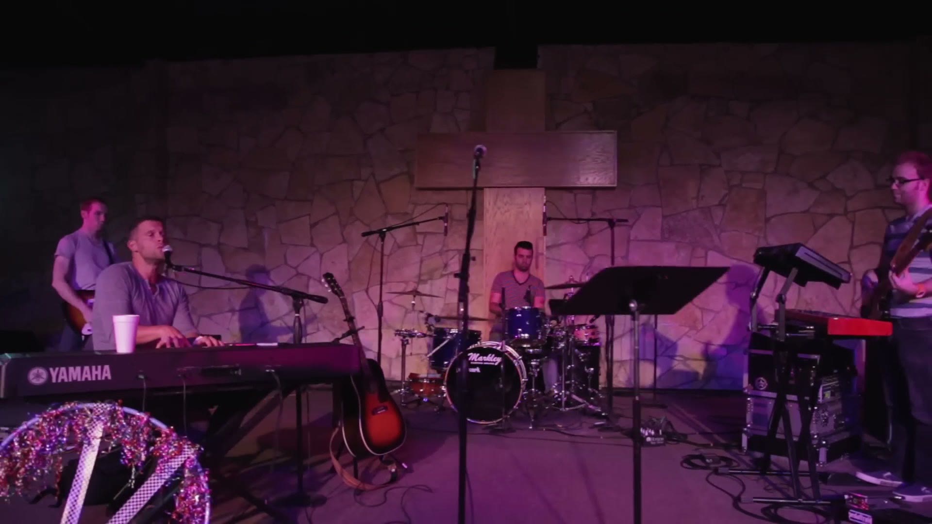 A worship band on a small stage with keyboard, drums and guitars, lit in purple against a stone wall