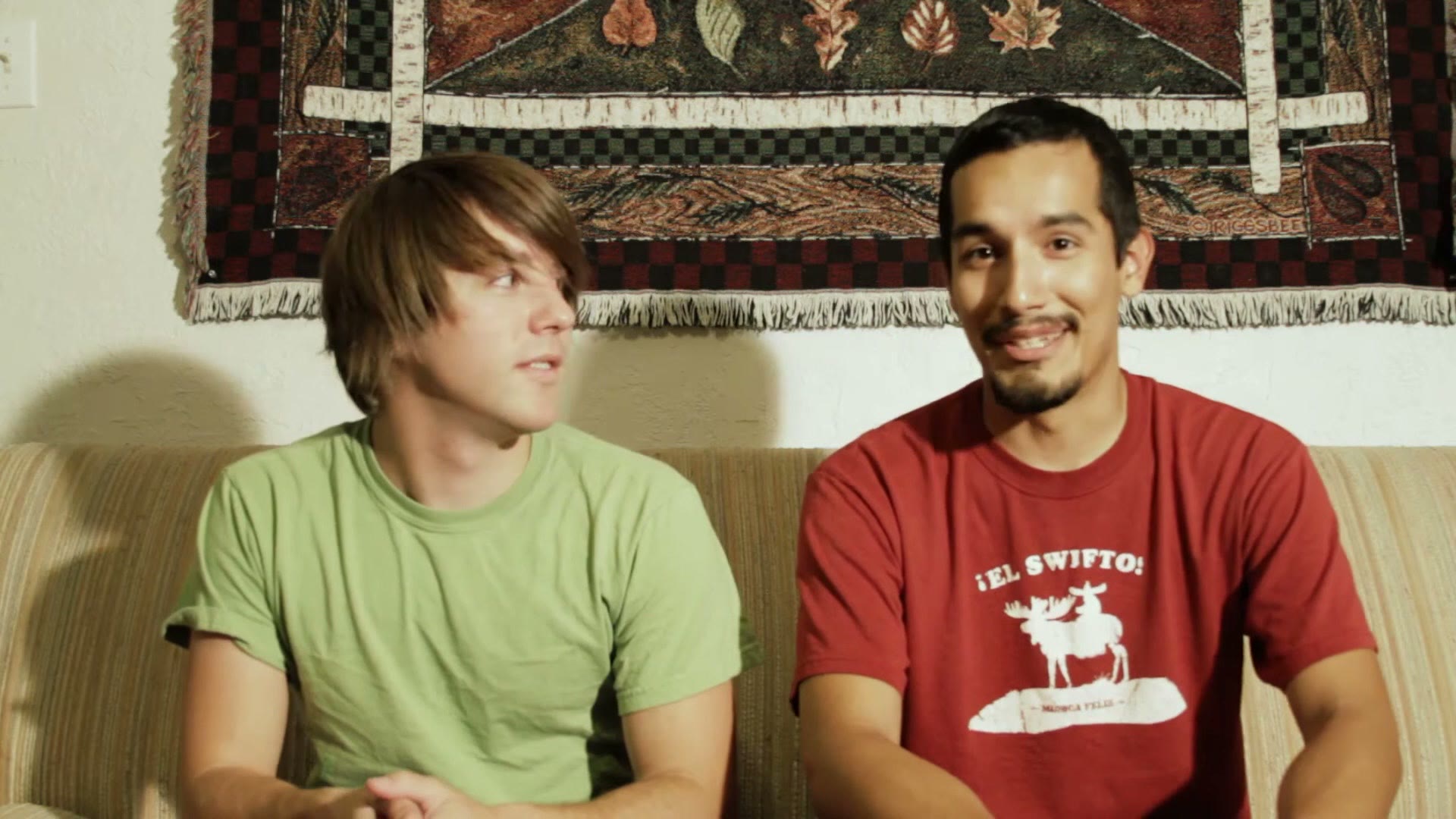 Two college students sitting on a couch talking to camera, the one on the right wearing a red moose graphic t-shirt