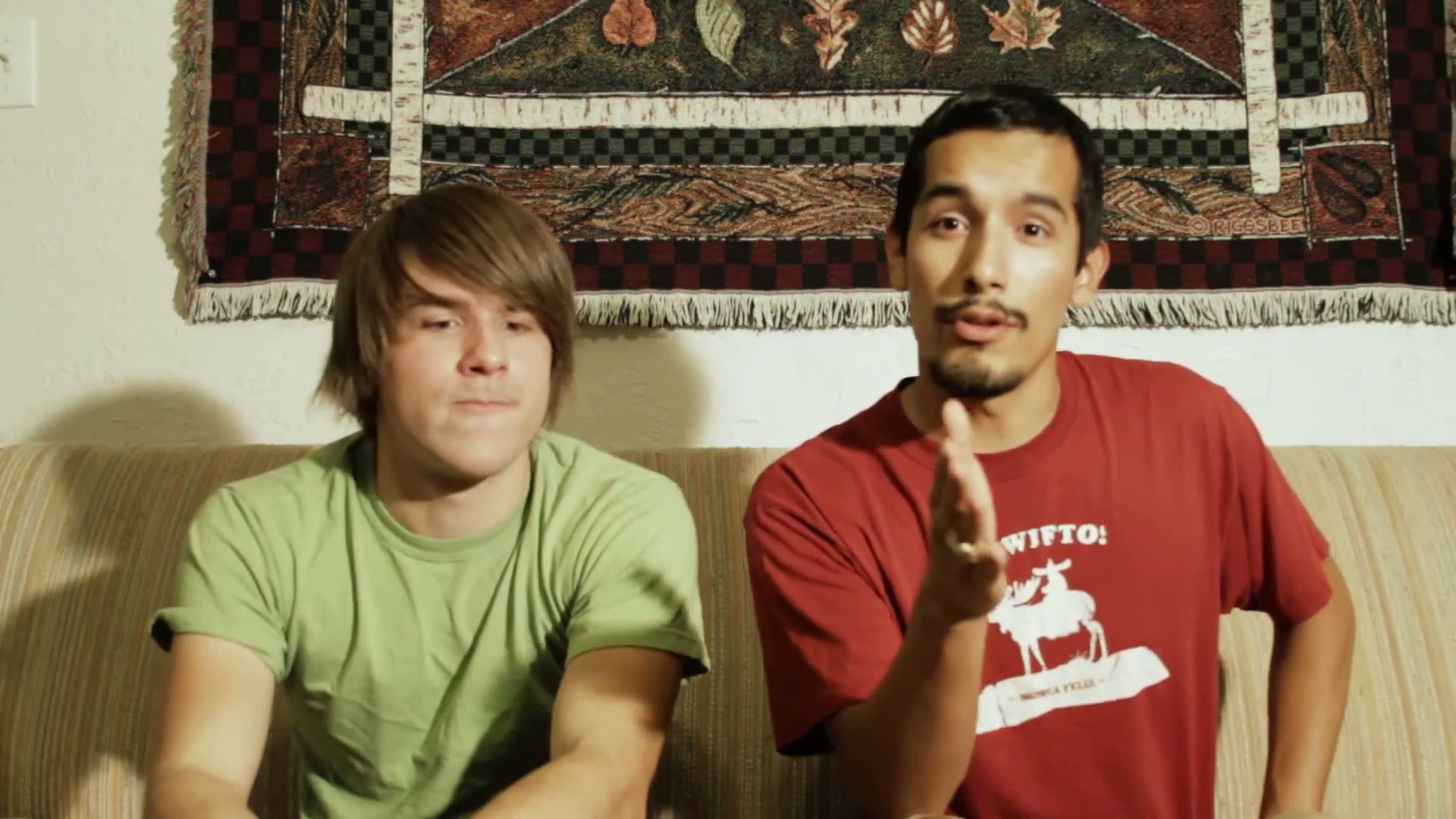 Close view of the two students on the couch, the one on the right gesturing as he explains something, with a mustache clearly visible