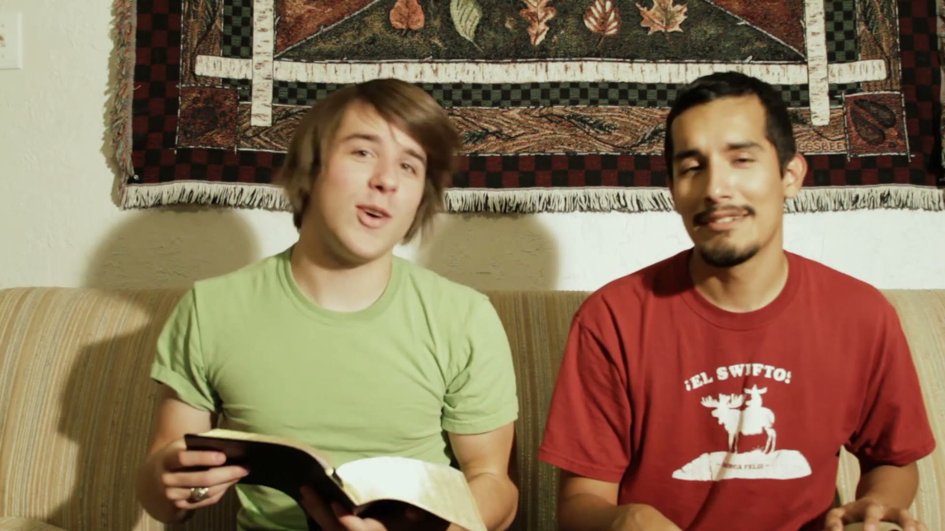 One of the students holding an open Bible on his lap while the other looks on from the couch
