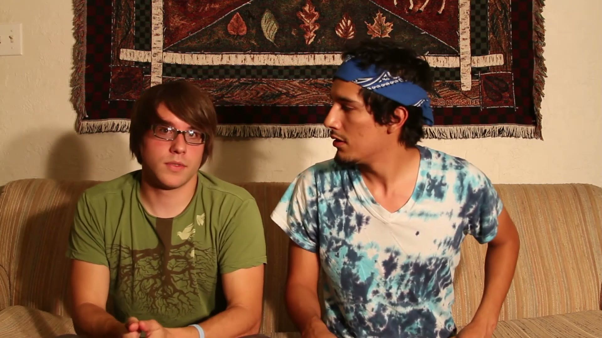 Two college students on a couch, one wearing glasses and a green shirt with a tree graphic, the other now wearing a tie dye shirt and a blue bandana